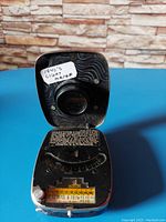 Front view of the 1940's vintage light meter with the hinged cover open, showing the circular sensor and calibration dial with numeric scale and instructions inside the lid.