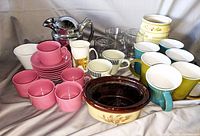 Assorted tea and coffee cups including pink matte cups with saucers, white cups with floral and striped designs, ribbed blue and yellow cups, cream ceramic serving dish, and glass pitcher with metal handle.