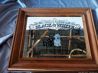 Framed mirror with Black and White Scotch Whisky branding on mirrored surface featuring dog images, set against a blue fabric background.