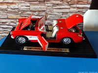 Front angled view of red 1957 Chevrolet Corvette model showing open hood, left door, and trunk, mounted on black base with plaque.