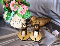 Photo showing woven basket with pink flowers, wooden duck, and two felt bird decorations arranged on a gray fabric background.
