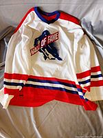 Front view of white hockey jersey with red, blue, and black stripes, Hall of Fame text and logo.
