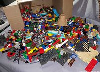 Large open cardboard box overflowing with a mix of colorful LEGO pieces including bricks, plates, pirate-themed parts, and terrain elements spread across a surface.