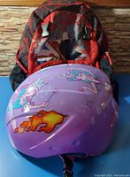 Kids backpack with dark red and black spider web and skull pattern shown with kids purple bike helmet placed on top.