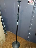 Adjustable black microphone stand with round base against a gray wall in a room corner.