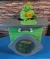 Front view of the green brick-styled piggy bank topped with Michelangelo figure and 'Awesome Earnings' window.