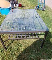 Outdoor backyard glass coffee table with clear glass top and metal lattice shelf under top viewed from an angle on grass