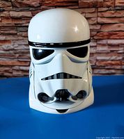 Front view of white plastic Stormtrooper helmet with black and gray details against blue surface and stone wall background.
