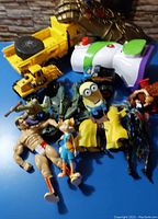 Mixed toy lot with Buzz Lightyear light-up glove, yellow dump truck, Minion figure, muscular action figure, and other assorted toy characters.