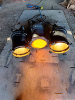 Three large stage lights positioned on ground; two emit white light, one emits orange light through a color filter; black metal body with barn doors