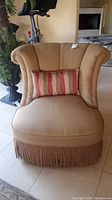 Frontal view of tan armless upholstered chair with decorative striped pillow and fringe skirt.