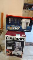 View showing Betty Crocker 5-speed hand mixer in box with attachments above the Cuisinart food processor box in kitchen setting.