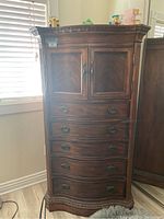 Front view of the tall wooden dresser with five lower drawers and two-door cabinet on top, showing carved molding and dark finish.