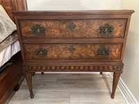 Frontal view showing two drawers with ornate metal handles and central key escutcheons. Detailed carved apron along the base and tapered, fluted legs.