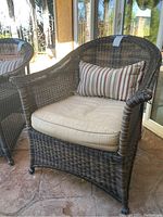 Photo of a single dark brown wicker patio chair with beige seat cushion and brown striped lumbar pillow on a stone floor patio in front of glass doors.