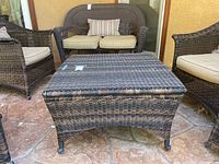 Angled side view of wicker patio coffee table showing woven wicker texture, curved legs with caster wheels, placed on tiled surface outdoors with matching wicker chairs and cushioned loveseat in background.