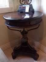 Front view showing the round wooden tabletop with drawer and the ornate metal base with wooden feet.