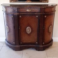 Front view of the vintage side table/credenza showing the central drawer, curved doors with embossed leather and ornate metal medallions.