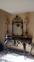 Full view of ornate black metal side/entry table with a mirror hung above and lamps on the table in a room corner, showing style and scale.