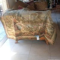 Woven tapestry depicting a pastoral scene with figures and nature, draped over furniture, showing color and design details.