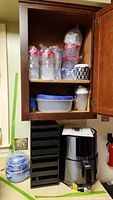 Photo of Shaq air fryer, Dash egg cooker, plastic food containers, disposable cups, plates and ceramic mug inside wooden kitchen cabinet.