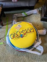 Disassembled bar stool showing round yellow seat cushion with PAC-MAN logo and chrome tubular frame pieces wrapped in protective material on carpeted floor.