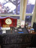 Full view of the lot showing the table clock, crystal heart, butterstone, metal blades, message tin, and other miscellaneous small items on a black wicker cabinet beneath a window.