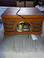 Front view of the Crosley stereo turntable showing wooden cabinet, metal radio dial, volume knob, speaker grills, and power cord laid on bed.
