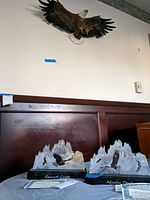 Wide shot showing two limited edition eagle sculptures labeled 'Summit Lords' and 'Mystic Peak' on a cloth-covered surface with an eagle wall hanging mounted on the wall above.