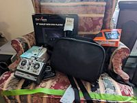 Photo showing Envizen 9" tablet with DVD player boxed, HP Sprocket printer, RCA DVD player, black case and black RC controller transmitter on patterned fabric chair.