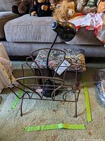Footstool with needlepoint cushion, wire magazine rack in front, and black goose neck lamp behind on carpeted floor.