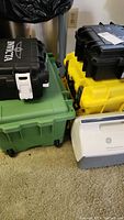 Stack of various colored plastic boxes including black Invicta watch boxes with white latches and handles, sitting on a green Igloo cooler.