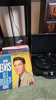 Record player closed case with stack of LP records in front, showing Elvis G.I. Blues album cover and others behind.