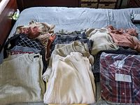 Overview of folded men's sweaters and shirts in neutral and warm colors including sweaters with various knit textures and styles arranged on a bed.