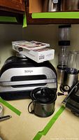 Ninja Foodi Grill, a blender with attachments and a small Crock-Pot on a kitchen counter with masking tape around items.