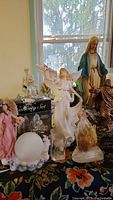 Photo showing a variety of religious statues and figurines including a large ceramic Mary statue with blue cloak, several angels including one holding a book, and a glass nativity set in a box.