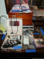Photo showing three wooden jewelry boxes on a table with assorted jewelry including boxed costume rings, silver chains, and some packaged jewelry pieces. Blue tape labels visible.