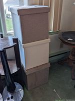 Photo shows three fabric covered storage boxes stacked on each other in front of window, neutral colors grey, white, and brown. Boxes are cubic shaped with tufted, padded lids. No visible damage except possible water stains on bottom box.