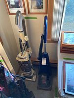 Photo showing Bissell Quick Steamer PowerBrush Pro carpet cleaner upright in blue and Shark Navigator upright vacuum in cream with dust bin, both next to a window and wall with framed photos.