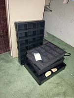 Three black watch display cases with individual square compartments and padded inserts, arranged stacked and side by side, on green carpet and against a wall.