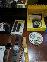 Overview photo showing multiple watches, boxed Invicta watch, S1 Sports Band box, metal watch bands and small watch parts on table surface.