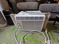 Full front view of Midea window air conditioning unit showing white plastic housing, front air vent slats, side window extension panels, and attached power cord with plug.