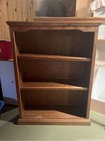 Front-facing photo of a wooden bookcase with three shelves, showing a medium brown wood finish and visible surface wear.