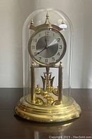 Front view of the anniversary clock under a clear glass dome showing the clock face and brass pendulum.