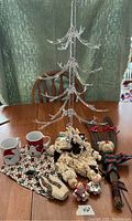 Clear plastic Christmas tree and assortment of Christmas items including mugs, plush lambs, small figurines, wooden Santa ornament, twig broom with bear and bow, and festive cloth