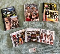 A photo showing eight compilation CDs laid out on a carpet, 3 in the top row and 5 in the bottom row. Titles visible include Rock Across Canada III, Women & Songs 7 and 8, Big Shiny Tunes 7, Classic Rock Gold, 80s British, and 103.9 Hawk Gold.