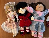 Three dolls: one porcelain head doll in a floral dress, one plush doll in a red coat and black beret, one plush doll in pastel outfit and slippers