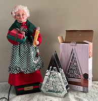 Photo shows full view of Mrs. Claus holding gift boxes and candle, with base labeled 'Noel'. Beside her is boxed mirrored triangular candle holder with Christmas message cutouts.