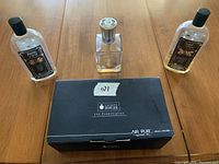 Clear glass oil lamp with silver burner cap next to black box and three partial bottles of lamp oil on wooden table.