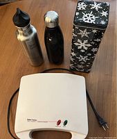 Photo of electric sandwich maker, two Kleen Kanteen bottles (one silver with spout, one black with metal lid), and black snowflake insulated drink carrier